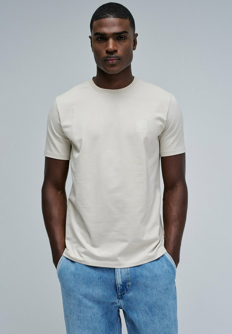 Camiseta de tejido de algodón suave, de manga corta y color beige claro, con un sutil logo en el pecho, combinada con jeans de mezclilla azul claro.