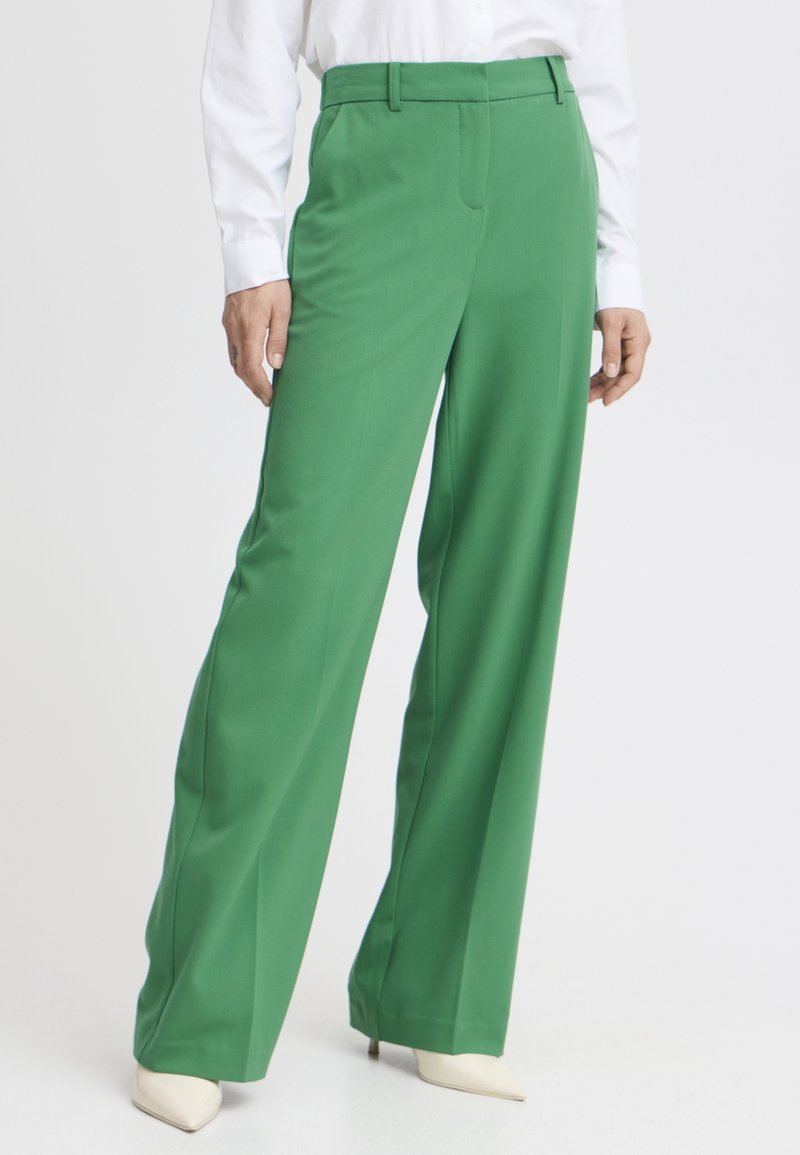 Femme portant un pantalon vert taille haute à jambes larges, associé à des chaussures blanches pointues et une chemise blanche à manches longues, debout sur fond blanc.