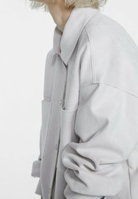 Giacca in suede grigio chiaro con colletto, tasche frontali e chiusura a bottone. Presenta una texture liscia e maniche leggermente oversize.