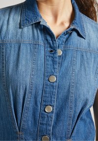 Žena v bezramenných modrých džínových šatech s kovovými cvočky na přední straně a límečkem.