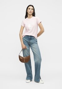 Γυναίκα με ανοιχτό ροζ μπλουζάκι και φαρδιά μπλε τζιν, κρατώντας μία καφέ τσάντα και φορώντας λευκά αθλητικά παπούτσια.