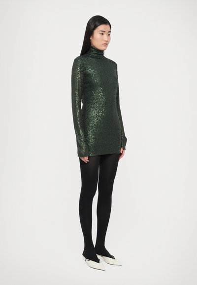 Zöld flitteres garbóruházat hosszú ujjakkal, fekete harisnyával és fehér hegyes orrú cipővel párosítva. Sima textúrájú, testhez álló kialakítás.