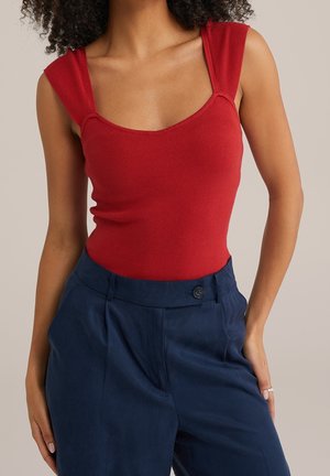 Červený žebrovaný body s hranatým dekoltem a širokými ramínky, přidaný k tmavě modrým kalhotám vysokého pasu s jediným knoflíkem a sklady.