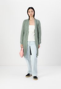 Ljusgrön öppen cardigan i ribbat tyg, bärs över en vit topp, ihop med ljusblå jeans och svarta sneakers, håller en rosa väska.