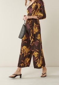 Mono marrón con hombros al aire y patrón floral amarillo, cintura ceñida y perneras anchas. Combinado con un bolso negro texturizado y sandalias.