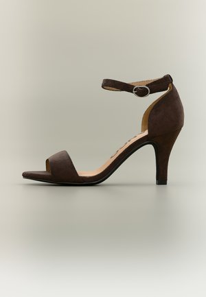 Hnědý semišový sandál na vysokém podpatku s otevřenou špičkou, kotníkovým páskem a stříbrnou přezkou, zobrazený z boku na neutrálním pozadí.