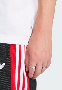 Biele bavlnené tričko s malou logo nášivkou, v kombinácii s čiernymi šortkami s červenými a bielymi pruhmi a textúrovaným vzorom. Na ruke strieborné prstene.