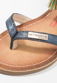 Zblízka hnědé sandály s třpytivým černým pásem přes prsty a kovovou destičkou s logem "Les Tropeziennes".