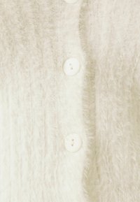 Texturerad vit kofta med mjukt, lurvigt stickat tyg, vertikal ribbning och fem stora vita knappar som stängningsdetaljer.