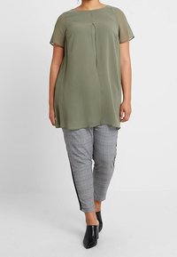 Olijfgroene, kortomse tuniek met een voorplooien, gecombineerd met grijze geblokte broek met zwarte zijstrepen en zwarte hakken.
