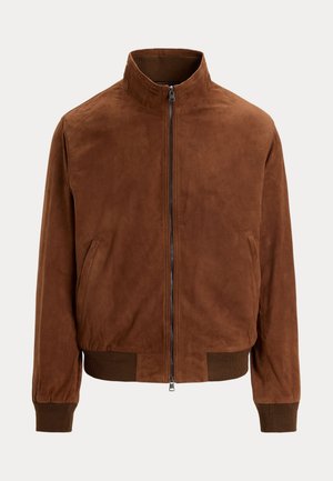 Νέος άνδρας που φορά καφέ μπουφάν, πράσινο πλεκτό πουλόβερ, λευκό πλισέ παντελόνι, καφέ μοκασίνια και γυαλιά ηλίου κρεμασμένα από το γιακά.