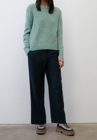 Person, die einen hellgrünen, flauschigen Pullover, schwarze weite Hosen und beiges, klobiges Schuhwerk trägt, steht vor einer schlichten weißen Wand.