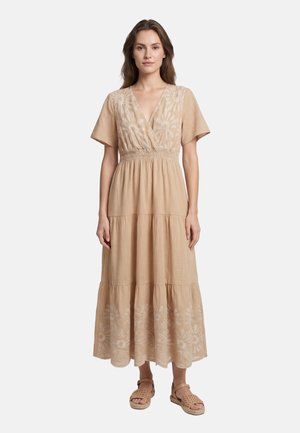 Donna che indossa un vestito midi beige a maniche corte con ricamo floreale e sandali intrecciati, in piedi davanti a uno sfondo bianco semplice.
