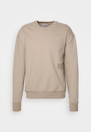 Sweatshirt beige avec un col rond, des manches longues et des poignets côtelés. Présente un logo discret en patch sur le bas gauche à l'avant. Texture lisse.