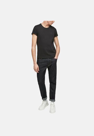 Mann trägt ein schwarzes T-Shirt mit hochgekrempelten Ärmeln, dunkle hochgekrempelte Jeans, weiße Sneaker, steht mit einer Hand in der Tasche und schaut zur Seite.
