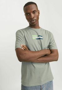 Szürke, rövid ujjú póló, amelyen egy színes "JACK & JONES" logó található a mellkason, puha pamut anyagból készült, laza fazonban.