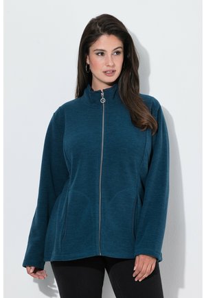 Vrouw met lang donker haar, gekleed in een teal ritsvest en zwarte broek, staand tegen een witte muur, kijkt iets opzij.