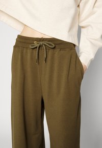 Olivgröna sweatpants med dragsko i midjan, med sidofickor och en mjuk textur. Tyget hänger löst runt benen.