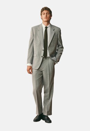 Junger Mann steht und trägt einen hellgrauen karierten Anzug, weißes Hemd, dunkle Krawatte und schwarze Loafers, mit einer Hand in der Tasche vor einem einfarbigen Hintergrund.