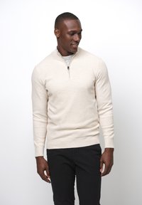 Maglione di maglia crema con colletto alto, chiusura lampo frontale e maniche lunghe. Abbinato a pantaloni neri, presentato su uno sfondo semplice.