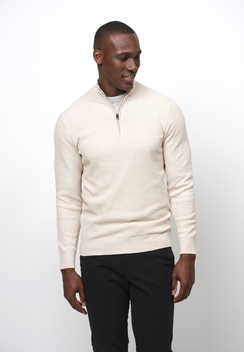 Maglione di maglia crema con colletto alto, chiusura lampo frontale e maniche lunghe. Abbinato a pantaloni neri, presentato su uno sfondo semplice.
