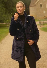 Jūras spēku krāsas mākslīgā kažokāda ar dubultā pārmantošanas dizainu, sudraba krāsas pogām, sānu kabatām un platu apkakli, valkāta virs melna krekla.