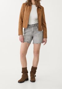 Personne portant une veste en daim beige, un haut blanc, un short en denim gris déchiré, et des bottes en daim marron qui arrivent au mollet, se tenant contre un fond blanc.