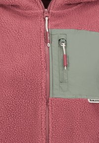 Nærbillede af en pink fuzzy jakke med en grøn lynlåslomme på brystet og matchende pink lynlåsgreb mærket "Sublevel."