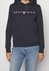 Granatowy hoodie wykonany z miękkiego materiału, z przednią kieszenią kangurką, z białym logo "GANT 1949" oraz wzorem herbu na klatce piersiowej.