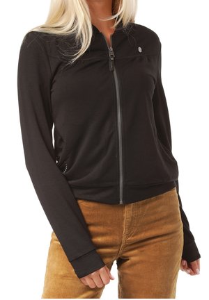 Woman wearing a black zip-up jacket with a small logo on the chest and brown corduroy pants, standing with hands relaxed by sides.