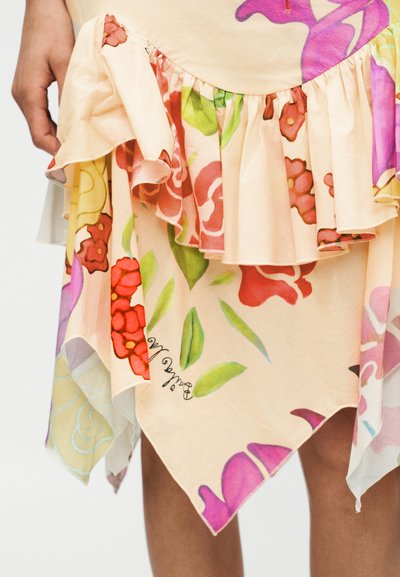 Φούστα με βολάν στο τελείωμα από ροδακινί ύφασμα με πολύχρωμα λουλουδάτα σχέδια σε κόκκινο, πράσινο, ροζ και μωβ, που φοριέται από άτομο με το χέρι χαλαρό.