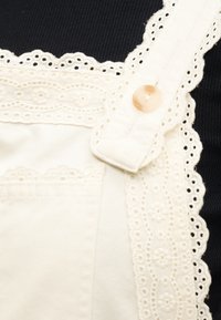 Λευκός ιμάντας ολόσωμης φόρμας με δαντέλα και κεντήματα λουλουδιών, με μπεζ κουμπί, πάνω σε ριγωτό μαύρο ύφασμα.