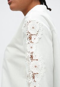 Λευκό πουκάμισο με περίτεχνη λευκή δαντέλα στο μανίκι, με λουλουδάτα σχέδια και απαλή υφή υφάσματος.