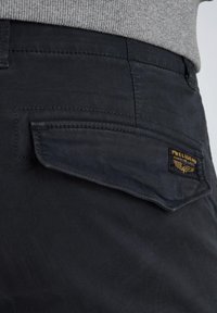 Dunkle Baumwollhosen mit texturierter Oberfläche. Verfügt über eine Klappentasche mit einem gelb-schwarzen Logo-Patch.