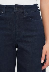 Sötétkék farmernadrág sima textúrával, magas derékkal és klasszikus öt zsebes dizájnnal, elöl gombos záródással.