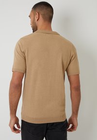 Krátký svetr s krátkým rukávem vyrobený z texturované pleteniny, s ribbovaným límcem a detaily na manžetách, nošený s tmavými kalhotami.