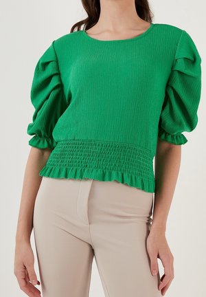 Mujer con una blusa verde brillante texturizada con mangas abullonadas y cintura fruncida, combinada con pantalones beige claro.