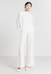 Weiße Bluse mit hohem Ausschnitt und drapiertem Vorderteil, kombiniert mit weit geschnittenen weißen Hosen und passenden spitzen Schuhen. Glatte Textur.