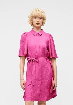 Kvinde med krøllet blondt hår iført en lyserød, tekstureret skjortekjole med korte pufærmer, krave og bindebælte i taljen.