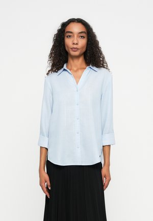 Light blue button-up shirt in soft fabric with a collar, long sleeves rolled at the cuffs, and a relaxed fit. Paired with a black pleated skirt.