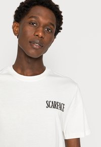White cotton t-shirt featuring "SCARFACE" printed in bold black font on the left chest, round neck, and short sleeves.