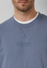 Muž oblečený v svetlomodrom crewneck svetri cez biele tričko, s textom "S.OLIVER ELEVATED SINCE 1989" na hrudi.