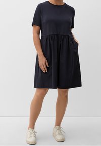 Robe bleu marine à manches courtes avec une taille froncée et des poches, fabriquée en tissu doux. Associée à des baskets blanches. Silhouette simple en ligne A.
