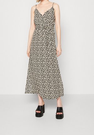 Femme portant une robe portefeuille noire sans manches avec des taches blanches irrégulières et des talons compensés noirs à bout ouvert, debout devant un fond uni.