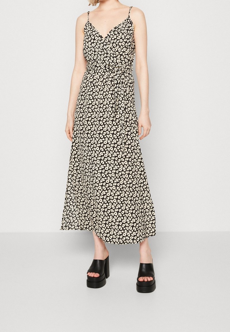 Femme portant une robe portefeuille noire sans manches avec des taches blanches irrégulières et des talons compensés noirs à bout ouvert, debout devant un fond uni.