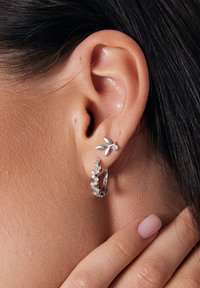 Srebrne kolczyki z motywem liścia, z małym akcentem diamentowym oraz teksturowanym kółkiem ozdobionym wieloma małymi, błyszczącymi kamieniami.