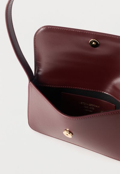 Ανοικτή μπορντό δερμάτινη τσάντα με λεπτό λουρί, χρυσό κουμπί κλικ και εσωτερική θήκη καρτών με την ετικέτα Victoria Beckham.