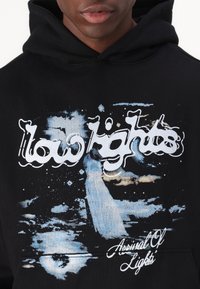 Svart hettegenser med et fargerikt grafisk trykk med abstrakte design, tekst som sier "lavbelysning," og frasen "Arsenal Of Lights."