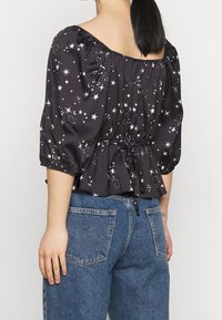 Blusa negra con estampado de estrellas, mangas abullonadas, ceñida en la cintura con un cordón y con un dobladillo en peplum. Llevada con jeans de mezclilla azules.