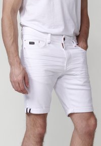 Pantalones cortos de mezclilla blanca con dobladillos enrollados, que cuentan con una franja de acento roja y azul. Diseño clásico de cinco bolsillos con una etiqueta de marca en la cinturilla.
